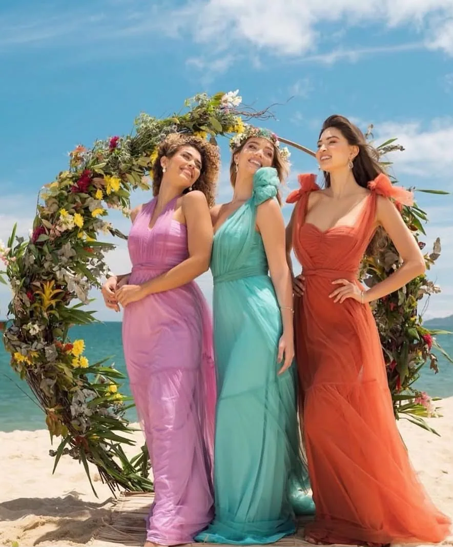 Madrinhas em rosa, verde água e terracota sob arco de flores na praia